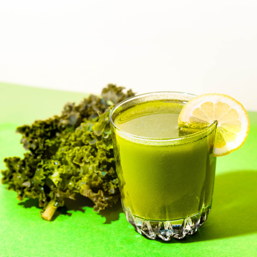 a glass of greens immunity tonic garnished with a slice of lemon on top and kale on the side on top of a green table