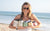 Image of a woman with shades carrying 5 bottles of protein shakes in her arms at the beach