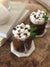 top view of two coffee mugs filled with chocolate drinks topped with mallows and choco bits with mint on the side on top of hexagon shape saucer.
