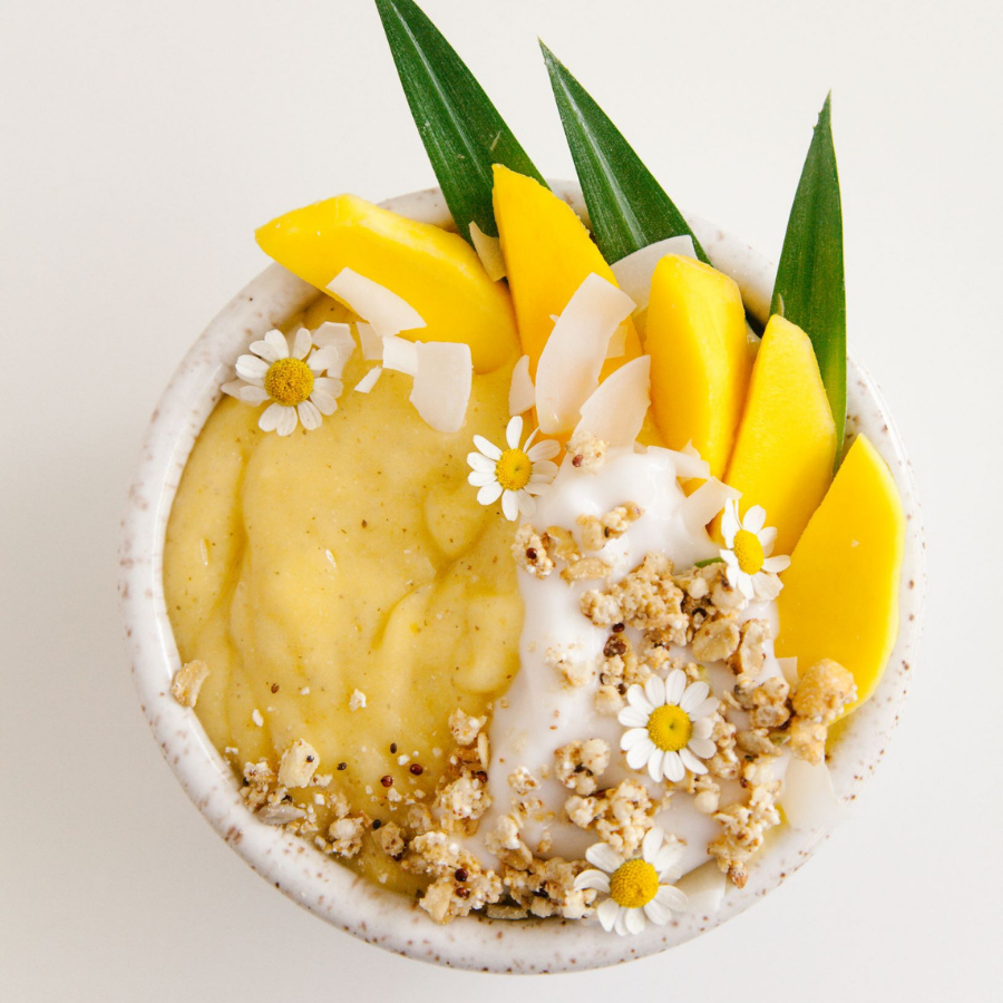 top view of mango pineapple chia smoothie bowl topped with slices of mango, pandan leaves, small white flowers, rice flakes, and coconut cream