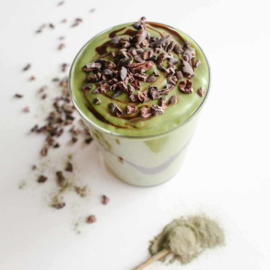 top view of a glass of organic shamrock shake topped with cacao nibs with scattered cacao nibs and organic pressed green powder with a wooden spoon on the side on the top of a white table