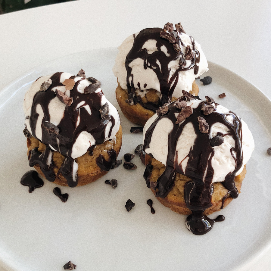 3 pumpkin protein muffins on a white plate topped with white coconut whip cream, drizzled with chocolate syrup, and sprinkled with chocolate crumbs.