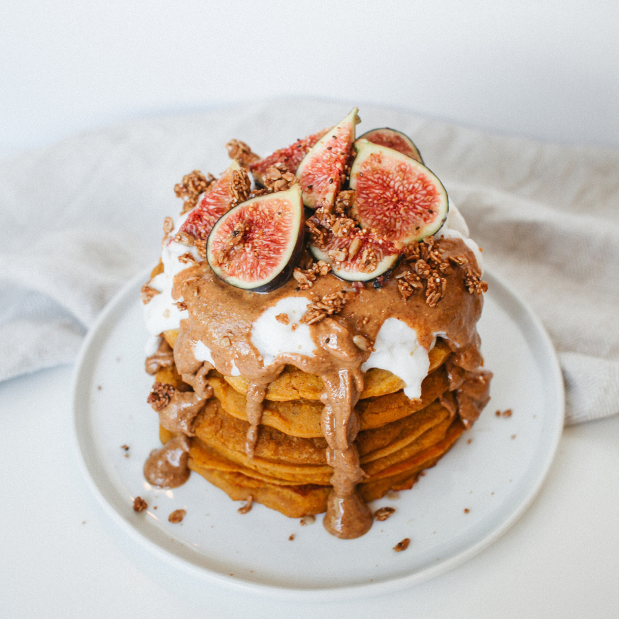 pumpkin pancakes on a white plate topped with peanut butter, sliced fruit, and sprinkled with pecan nuts. 