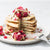 six-layered banana proteini pancakes with sliced portion topped with berries and squash seeds on top a plate surrounded by berries, squash seeds, and fork on the side