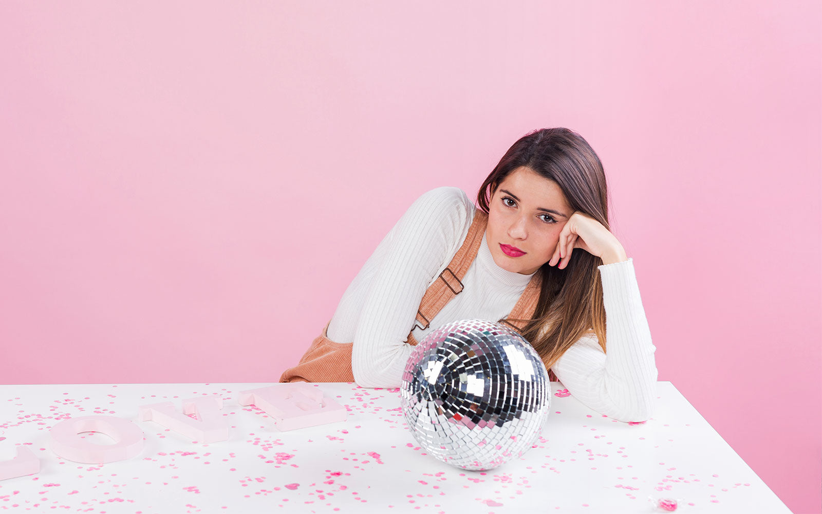 Image of a bored-looking woman leaning on a marble table top with a silver disco ball in front of her. Set against a pink wall.