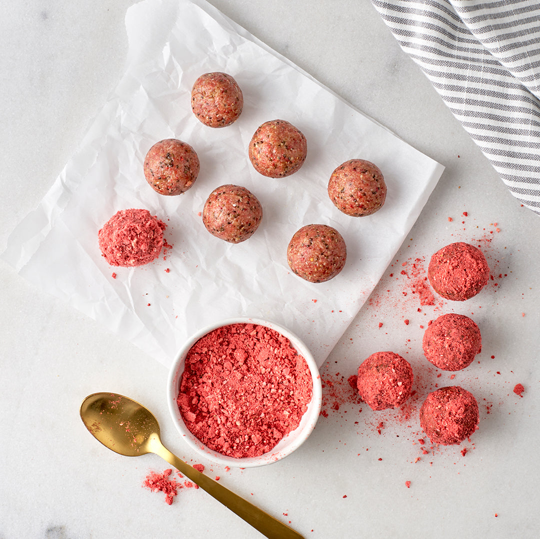 top view of a bowl of powdered red juice and a spoon on the side surrounded by a ball shape powdered juice on top of a parchment paper on top of a white table