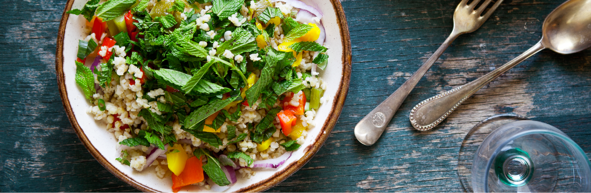 a bowl of salad with quinoa, mint leaves, cilantro, cherry tomatoes, diced red onion and mined garlic beside a glass and a spoon and fork. 