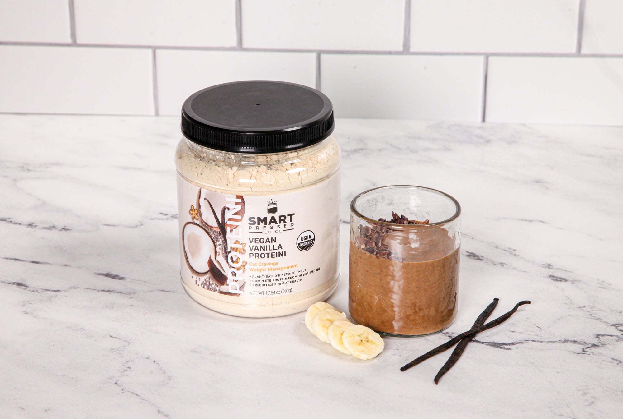 1 jar of Vegan Vanilla Proteini beside a glass of Arash's Cacao Proteini, slices of banana and 2 sticks of vanilla on a marble counter top.