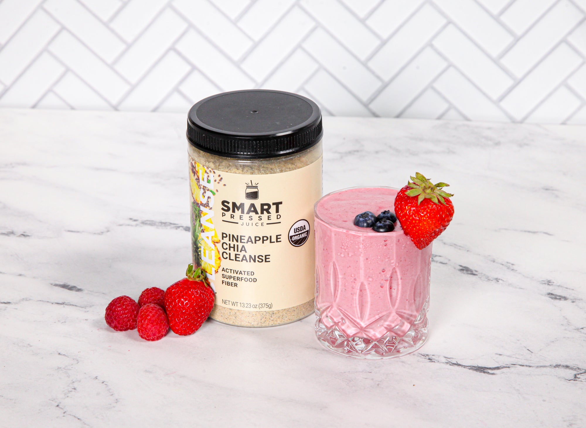 front view of a glass of pineapple chia cleanse signature smoothie topped with berries with a jar labeled as smart pressed pineapple chia cleanse with fresh berries on the side of the jar on top of a marble table