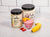 1 jar of Pineapple Chia Cleanse beside 1 jar of Vegan Vanilla Proteini and in front is a glass of Rose's Tropical Proteini topped with slices of apricot  beside a banana and slices of strawberry on a marble counter top.