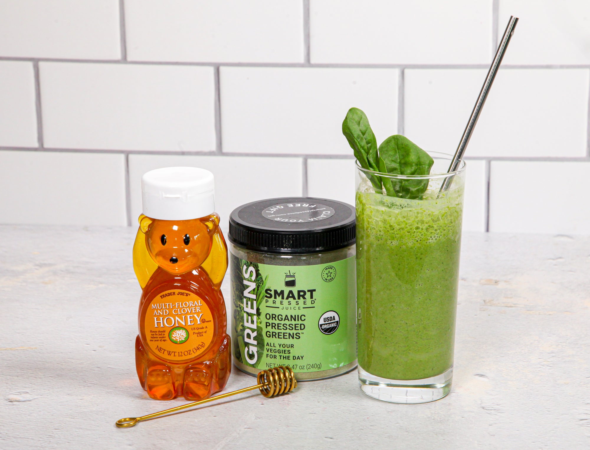 a glass of green apple greens smoothie, a jar of smart labeled organic pressed greens and a bear-shaped container filled with honey and a honey digger on the front on top of a white-painted table