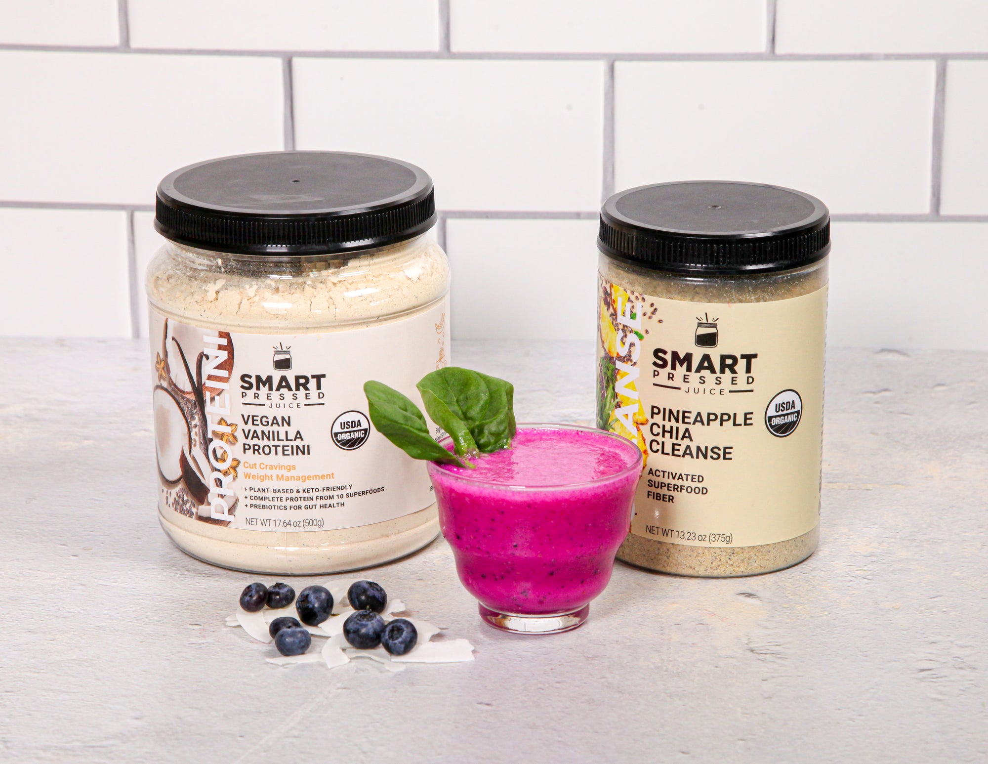 two smart labeled jar with a glass in the middle filled with dragonfruit cut cravings smoothie with garnish with two leaves of spinach surrounded by blueberries and coconut chips on top of a white table