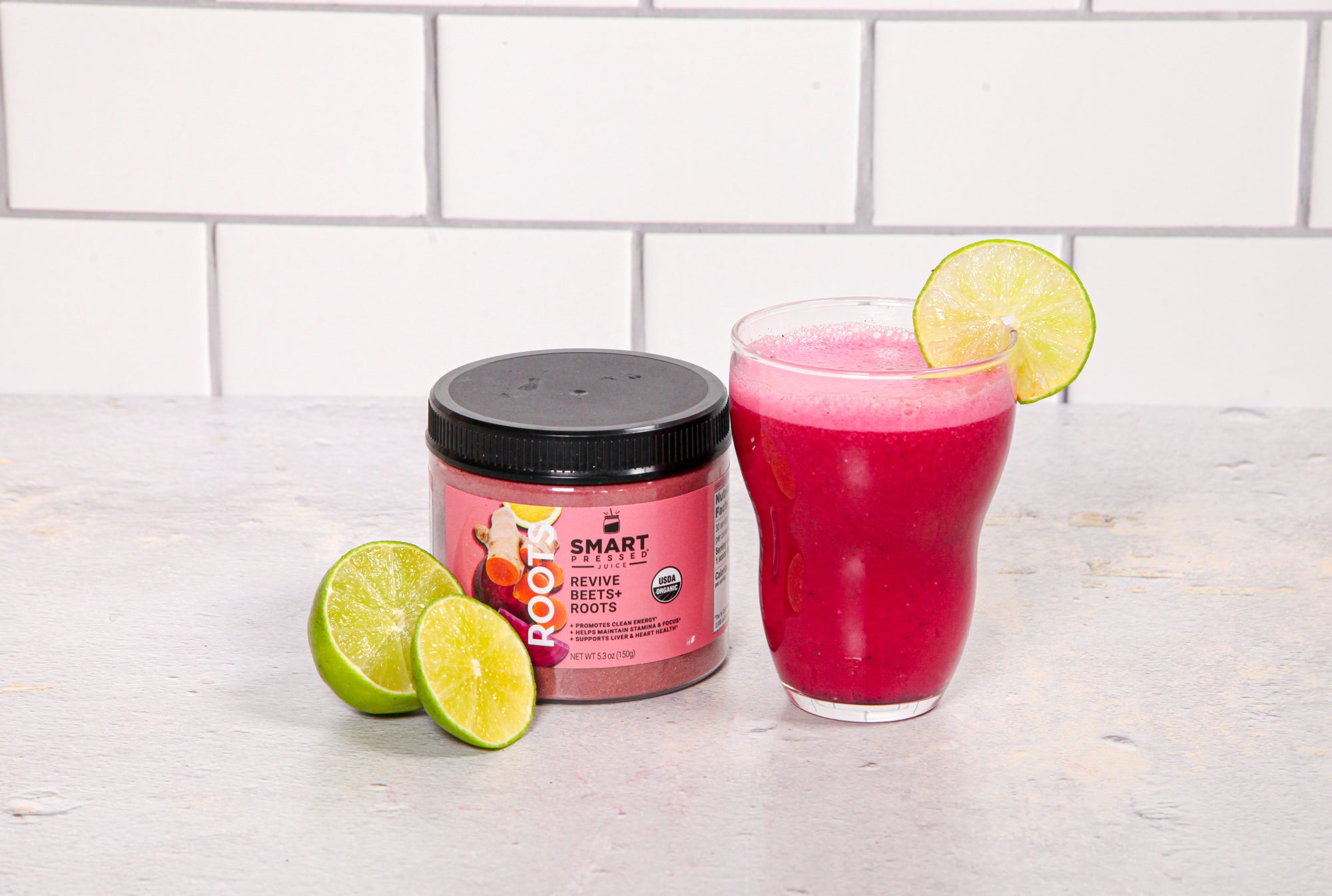 a glass of hibiscus beets refresher garnish with a slice of lime and a smart revive beetroots labeled jar and slices of lime on the side on top of a white table