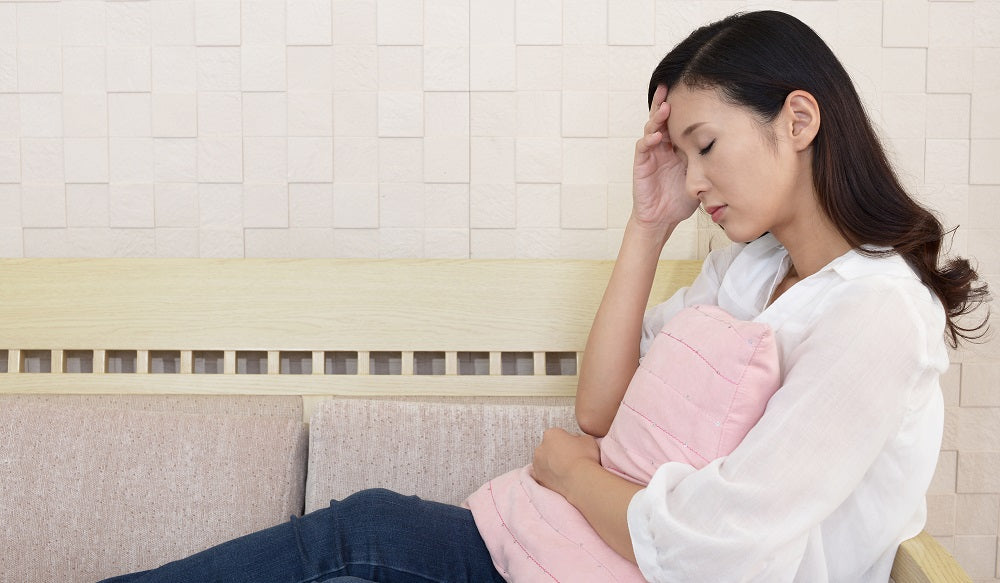 A sad woman sitting on a couch hugging a pillow with her left arm and holding her head with her right hand.