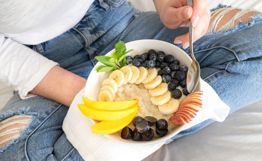 top view shot of a woman holding a plate of fruits with blueberries, sliced banana, sliced mango, black grapes, sliced apples on her right hand and holding a spoon on her left hand. 