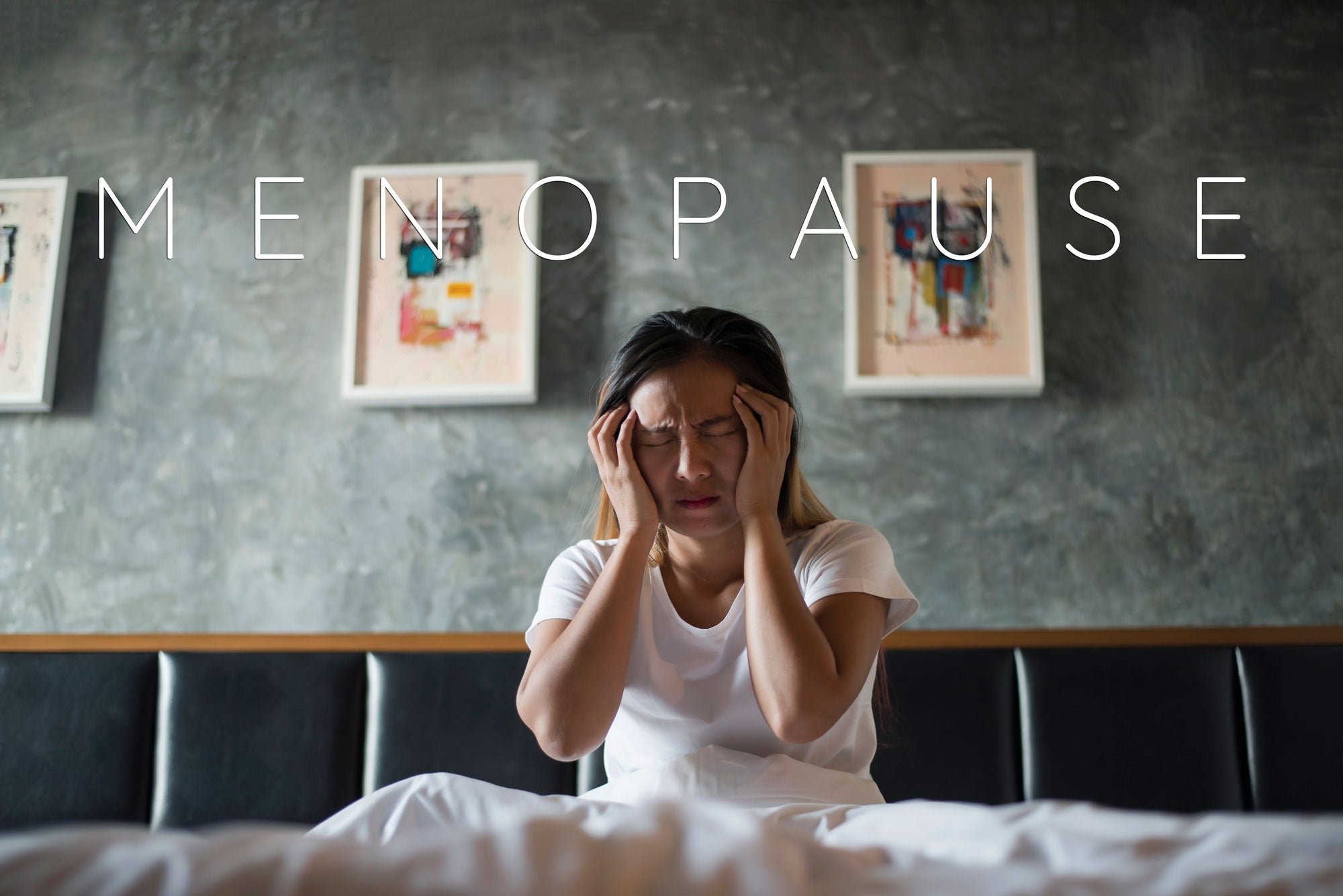 Image of a woman sitting on her bed in agony. Behind her is a grey wall with 2 art frames, and in the middle of the image is a text that says, "Menopause".