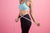 Image of a fit woman's body in a sports wear holding a measuring tape around her waist. She is standing against a pink wall.