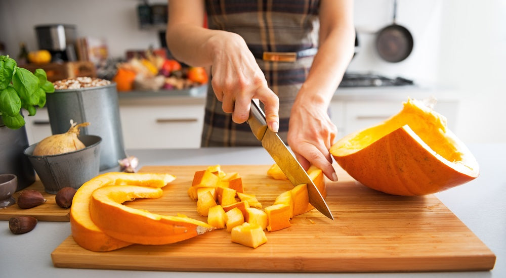 Image of a half-body woman in her kitchen chopping a yellow pumpkin. 
