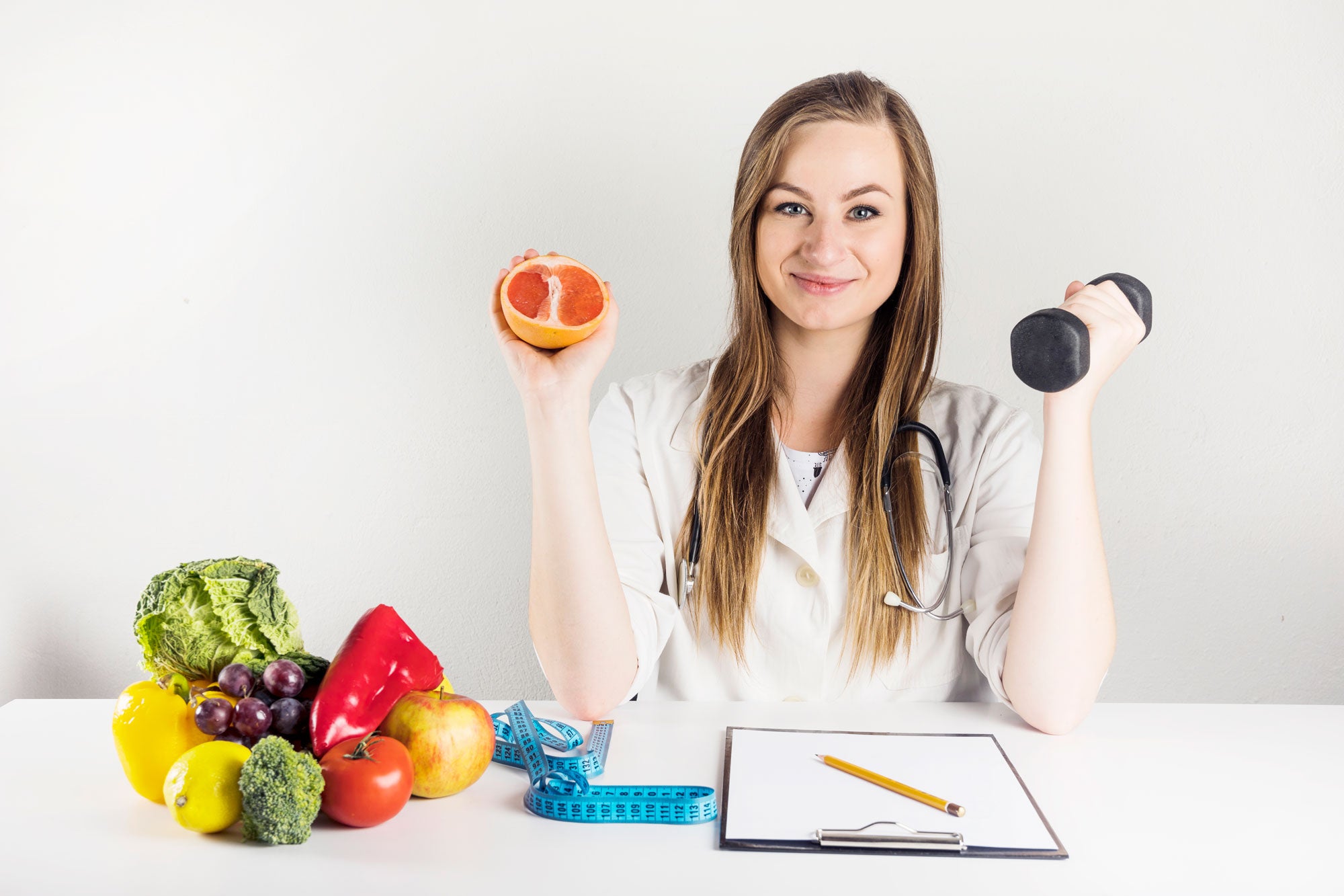 Image of a woman holding a grapefruit on her right hand and a dumbbell on her left hand. In front of her is a white table with a bowl full of fruits and vegetables and a clipboard with a paper and pencil.