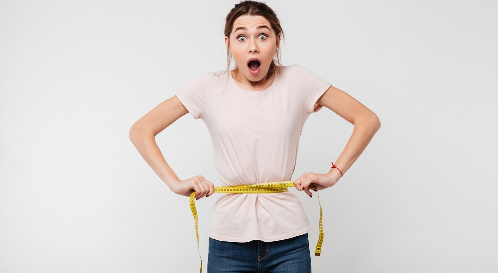 Image of a woman looking shock while holding a measuring tape tightly wrapped around her waist. She is standing against a white background.