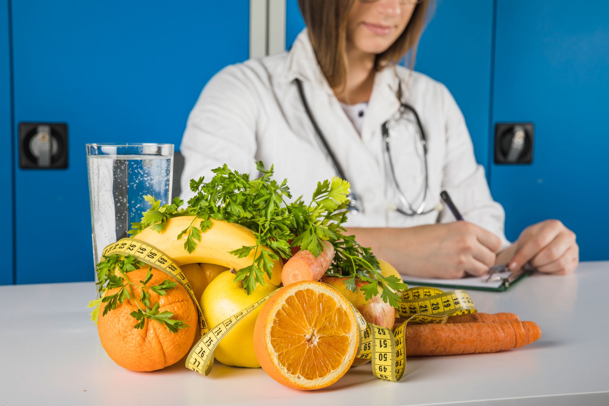 Image of a female Doctor writing in a paper on the white kitchen top. In front of her is a glass of water and fruits like oranges, banana, lemon, carrots, and vegetables like carrot and parsley with a measuring tape along with the foods. 