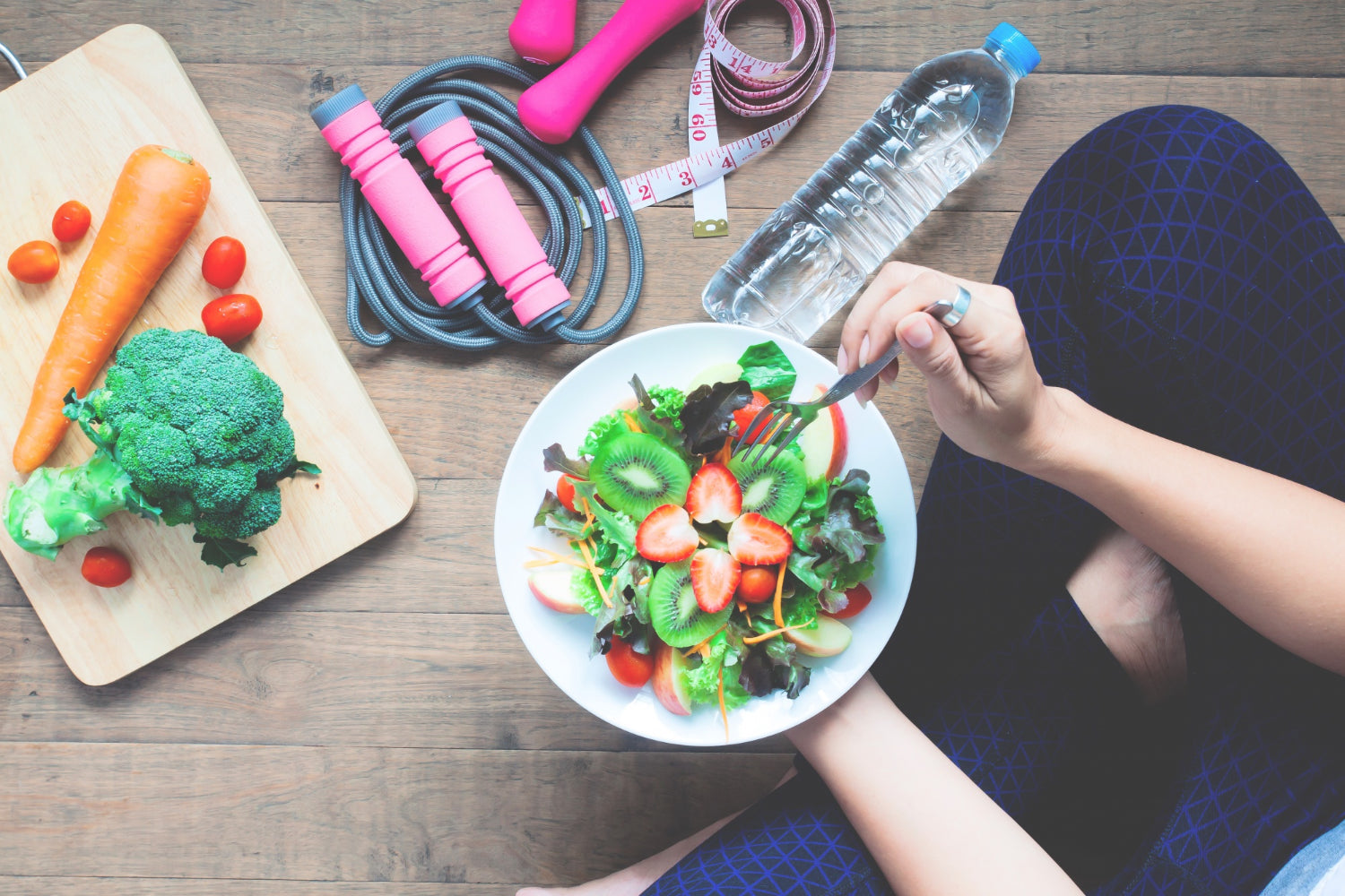 Top view of someone sitting cross legged and holding bowl of salad next to cutting board with vegetables, jump rope, measuring tape and bottle of water.