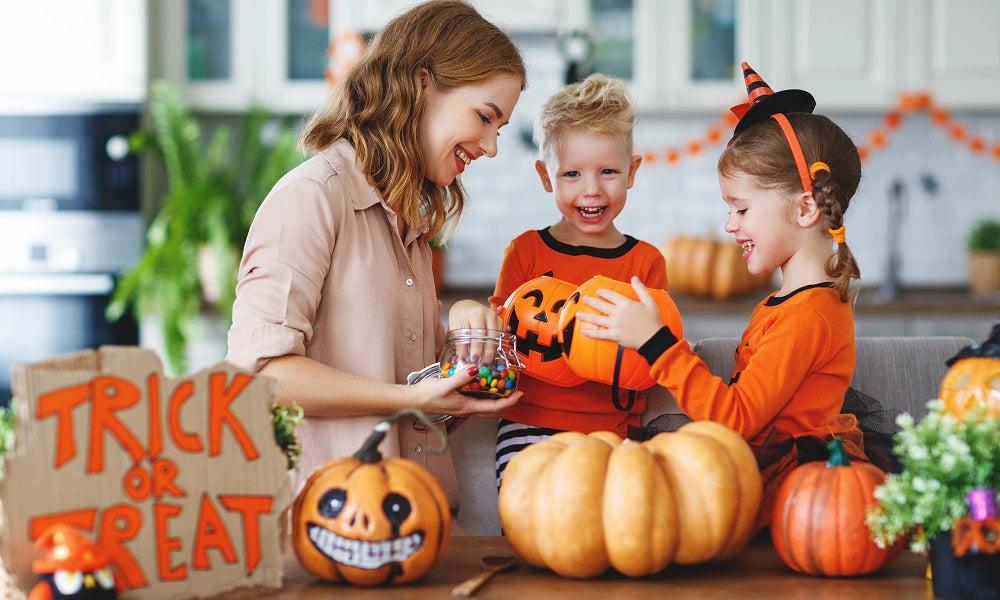 front view of a mother holding a jar of candy and a boy and a girl holding a pumpkin shape basket with a pumpkin curved as a skull and a two pumpkin with a wooden spoon on the side on top of a wooden table