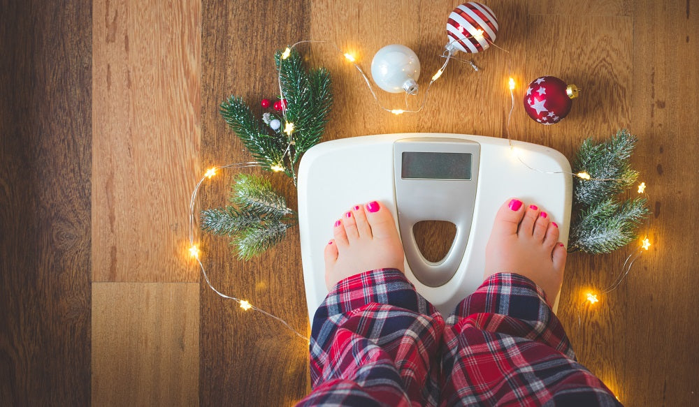 Top view image of a a female feet on a weighing scale with Christmas ornaments surrounding it on a wooden floor.