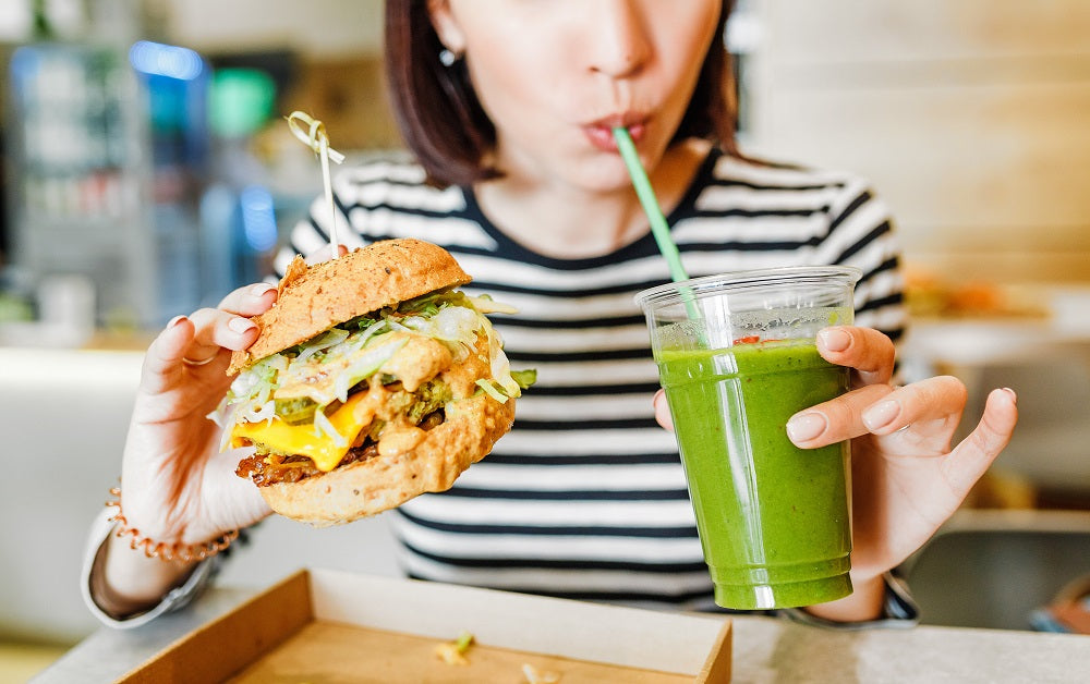 A woman sipping a glass of green smoothie while holding it with her left hand and holding a burger on her right hand.