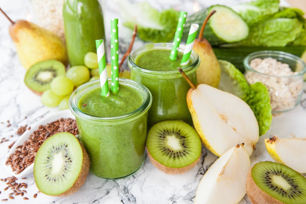 two glasses of green smoothies with two green patterned paper straws on top of the smoothie placed on top of a marble table. smoothie glasses are surrounded by cut kiwis, pears and grapes.