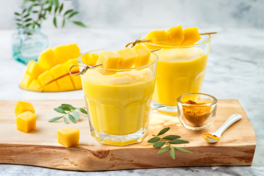 two glasses of mango turmeric smoothie with sliced mango on a stick on top of the glass surrounded by a small glass of turmeric and a white plastic spoon with turmeric powder and three cubes of sliced mango on the side placed on top of the wooden plank.