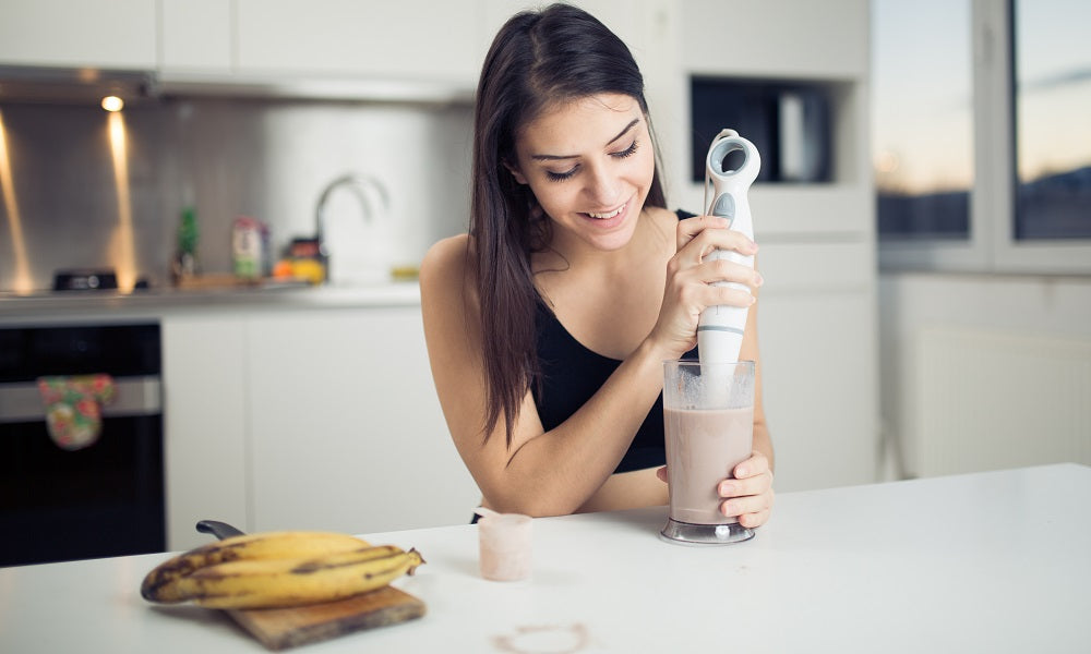 Image of a happy woman making her smoothie using a hand-held blender on her kitchen top.