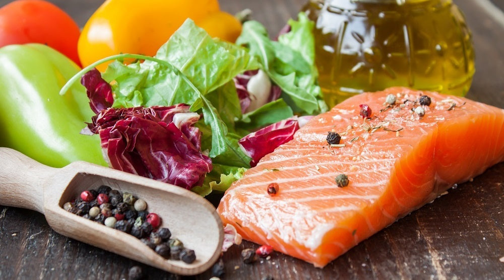 A slice of salmon topped with whole pepper corns, beside is a glass scoop of pepper corns, some green vegetables, a pouring glass of olive oil, and some yellow, red and green peppers side by side on a wooden surface.  