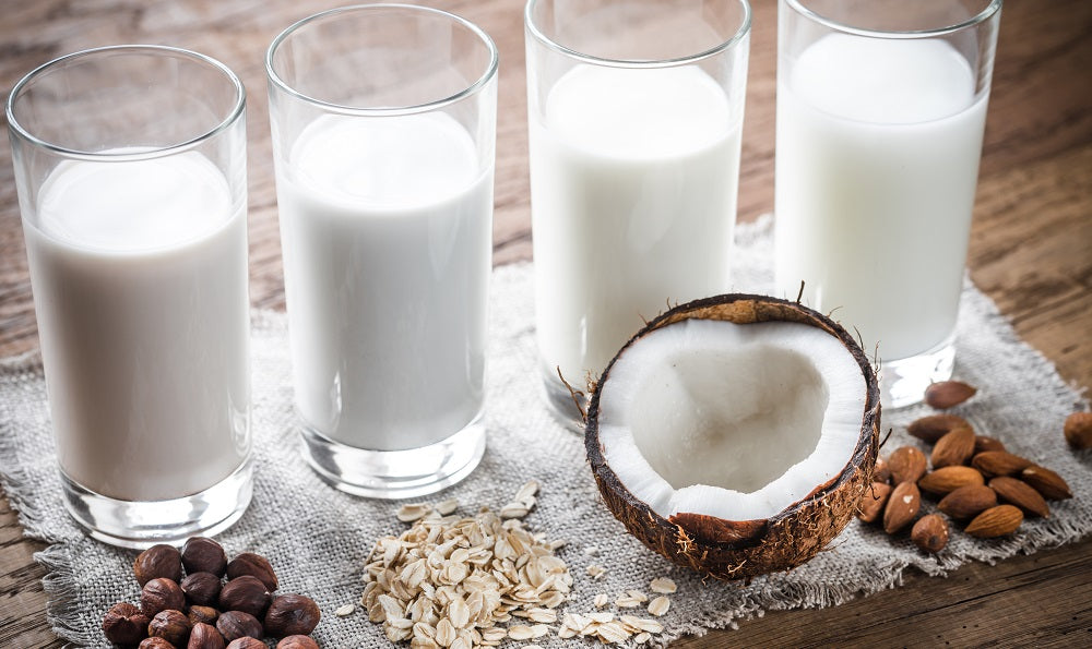 4 tall glasses of milk side by side- in front of the 1st glass is a handful of hazelnuts, 2nd glass is a handful of grains. 3rd glass is a half a coconut, and the 4th glass is a handful of almonds.