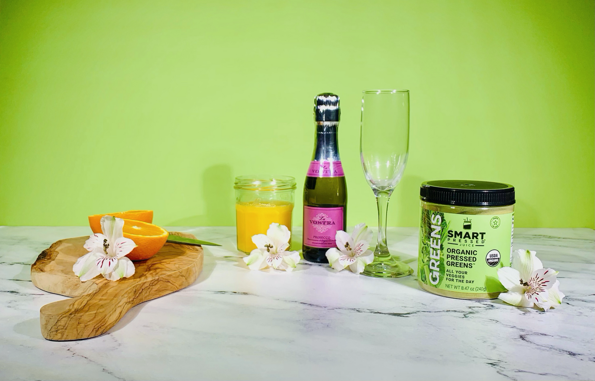 a cutting board with 2 slices of oranges and white orchid beside a glass of orange juice beside a bottle of mimosa, a champagne glass and a jar of Organic Pressed Greens surrounded by white orchid flower on a marble top against a green wall.
