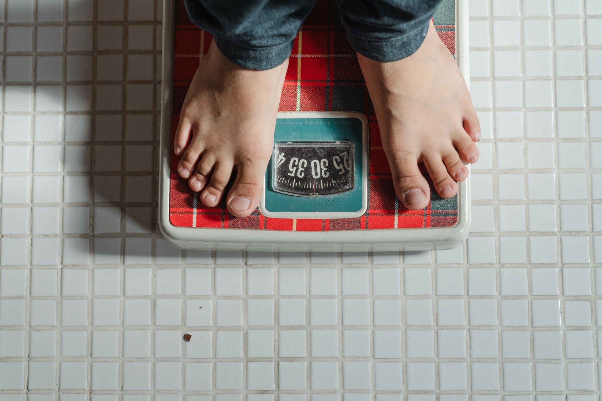 Image of a top view shot of a person's feet on a red weighing scale on a white tile flooring.
