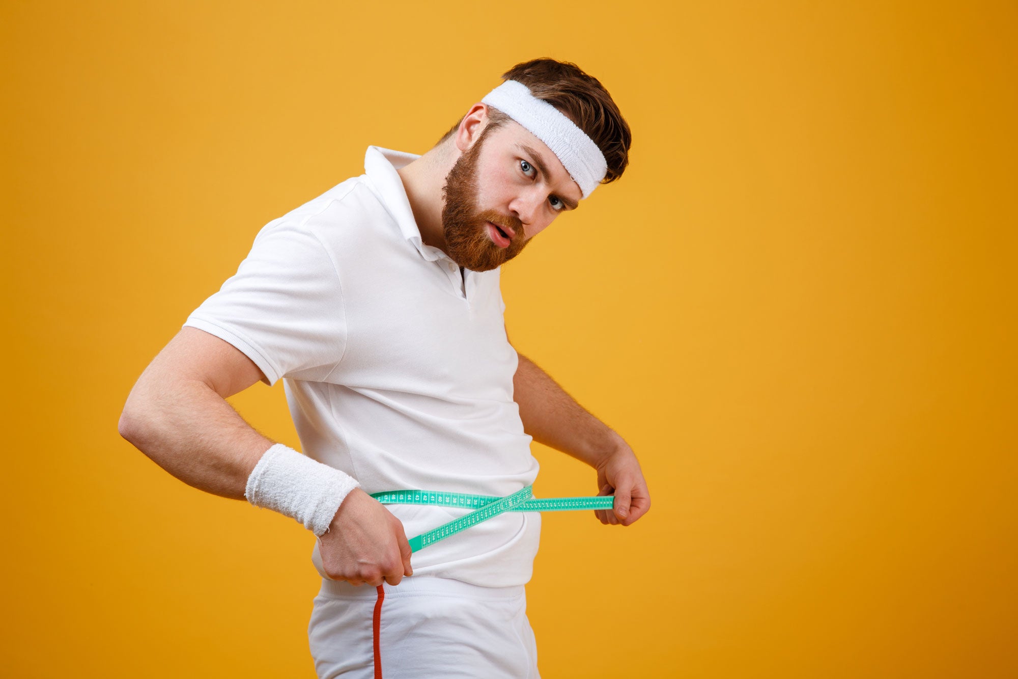 Image of a man in a white tennis wear holding a measuring tape around his waist. He is standing against a yellow wall.