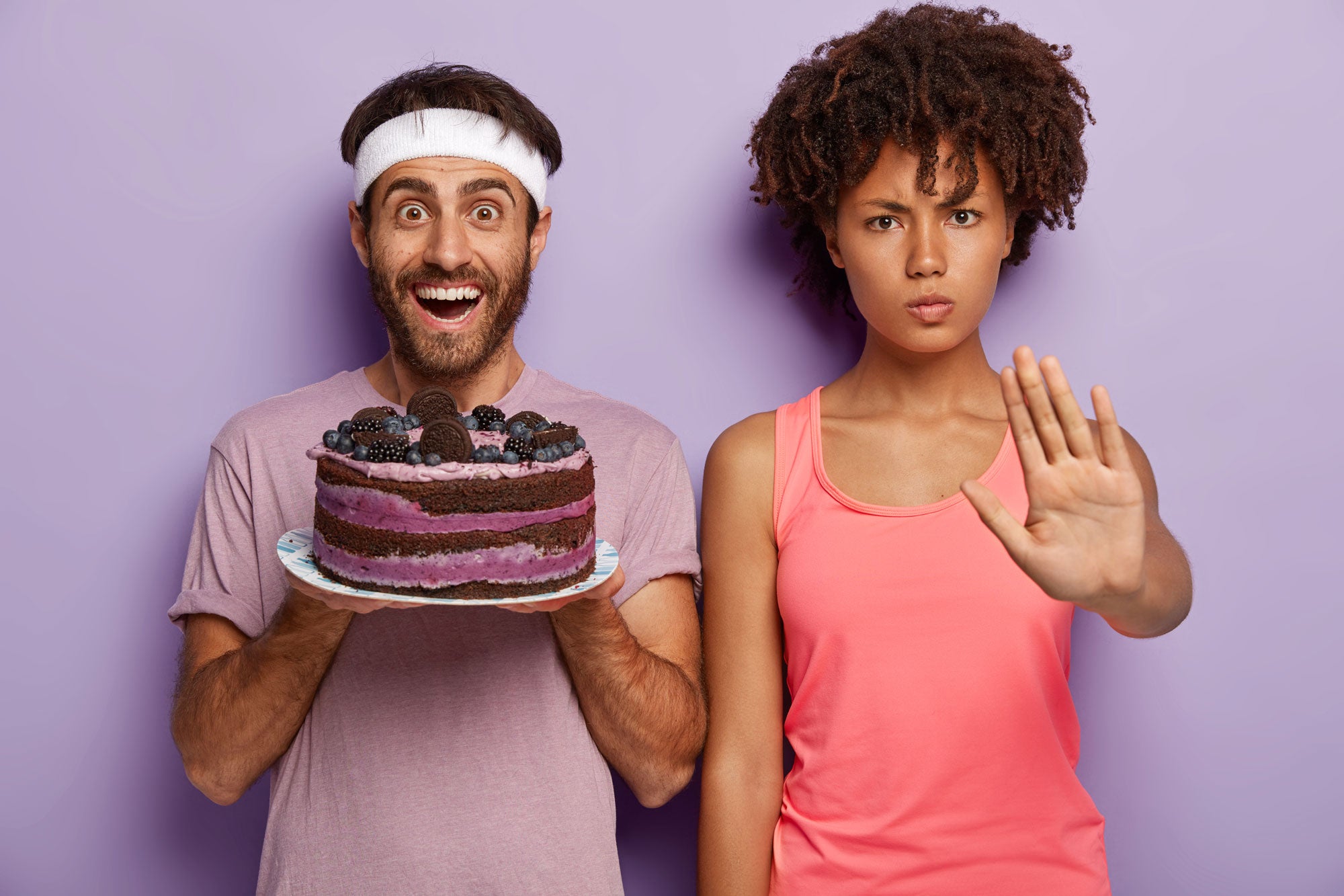 Image of a super thrilled man wearing a sports headband holding a 4-pound cake. Beside is a woman with a concerned face and a stop hand gesture. They are standing against a light purple background.