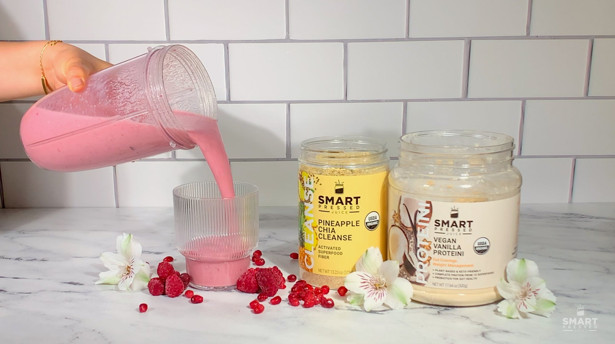 Pomegranate smoothie being poured into a glass with Pineapple Chia Cleanse and Vegan Vanilla Proteini next to the glass on a subway tile background