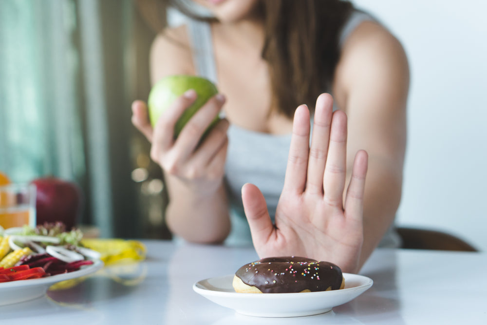 A woman holding a green apple on her right hand with a white saucer with chocolate donut sprinkled with nuts in front of her with her left hand on the side of the saucer on top of a marble table