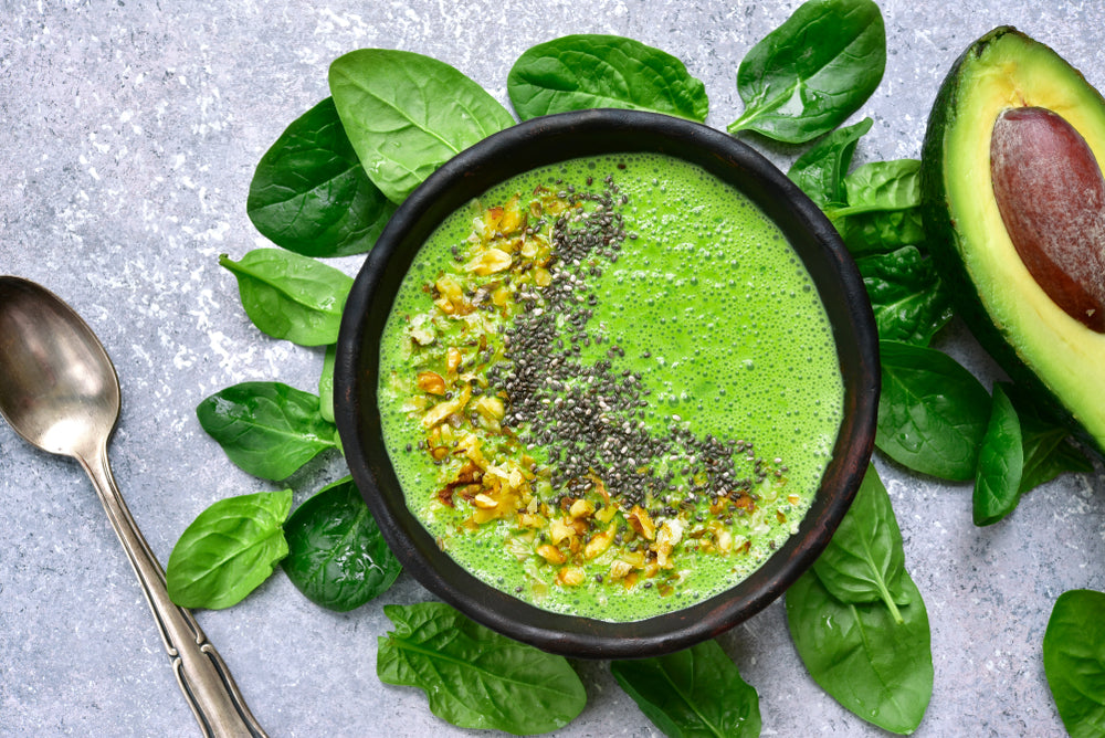 a bowl of green jalapeno pineapple smoothie sprinkled with chia seeds and nuts placed on top of a bunch of basil leaves with cut avocado and a spoon on the side