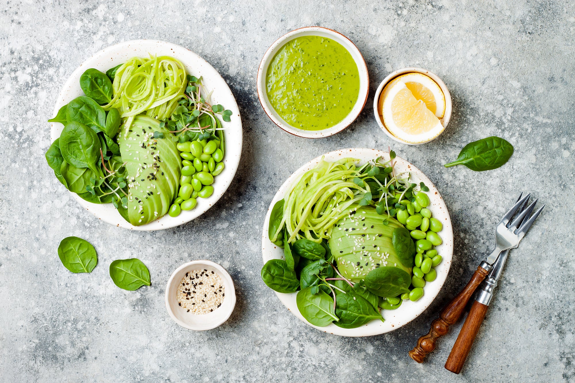 top view of two plates of fresh pressed greens salad dressing surrounded by two forks, a small bowl of slices of lemon, a bowl of pressed green salad dressing, and a small bowl of black and white sesame seeds on top of a concrete table