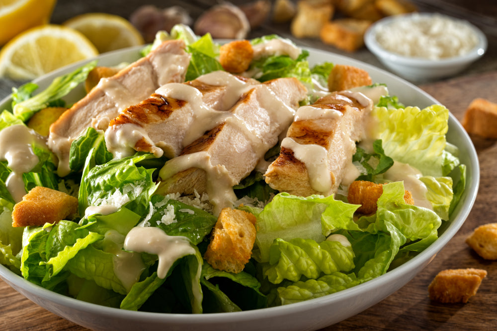 Chicken caesar salad drizzled with creamy dressing with croutons in a white bowl on a wooden table