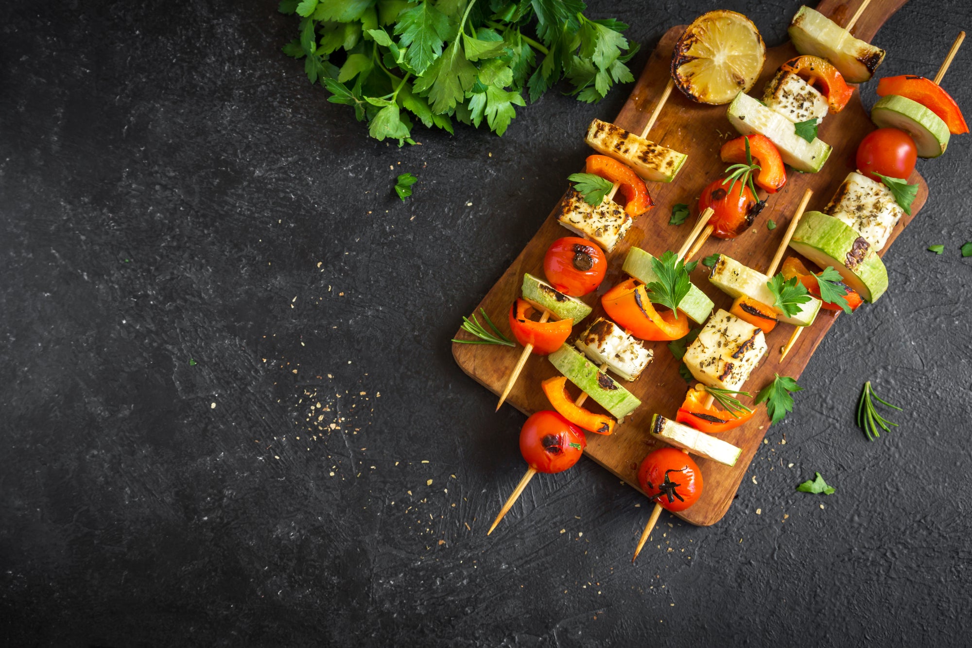 top view of veggie kebabs and a slice of lemon on top of a wooden chopping board with cilantro on the side on top of a black concrete table