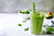 a glass of spinach avocado smoothie with chia seed and two green patterned paper straw surrounded with spinach leaves, a small white bowl full of spinach leaves, and a slice of avocado with seed