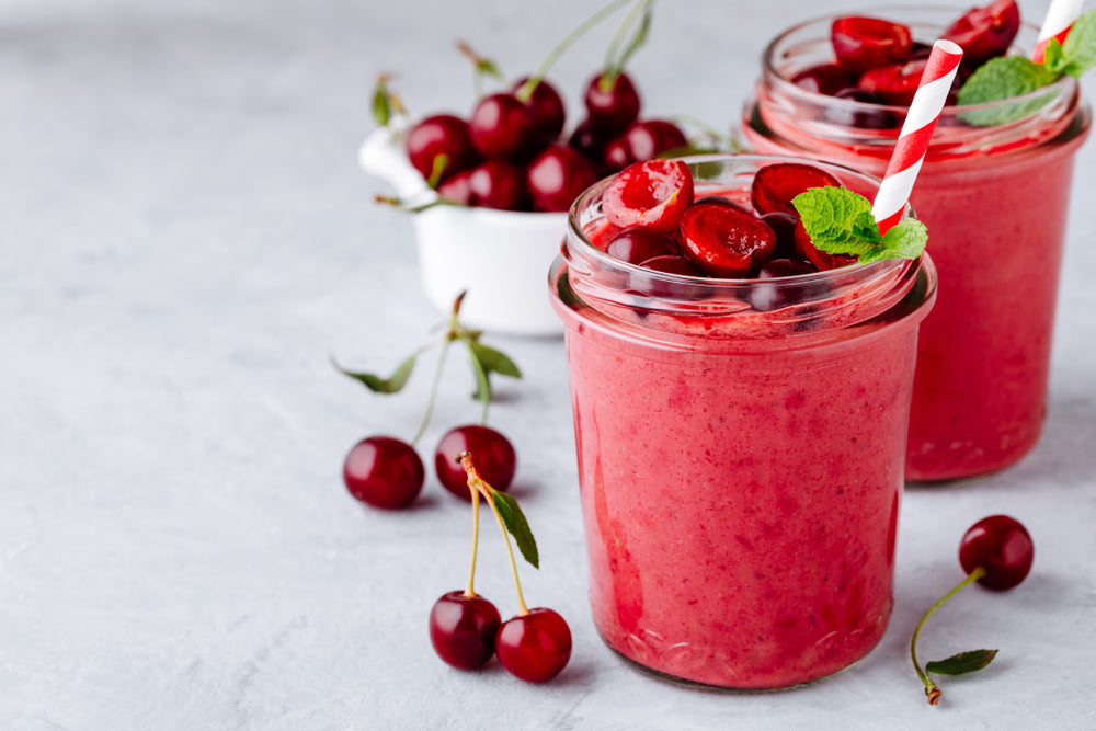 two glasses of beets smoothie topped with sliced cherries, mint leaves and red patterned paper straw and a white bowl of cherries and scattered cherries with twigs on it
