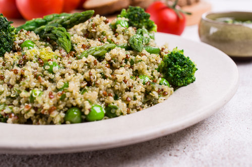 A plate of green tea quinoa and peas with roasted asparagus and steamed broccoli on the side on top of a white concrete surface with tomatoes behind the plate