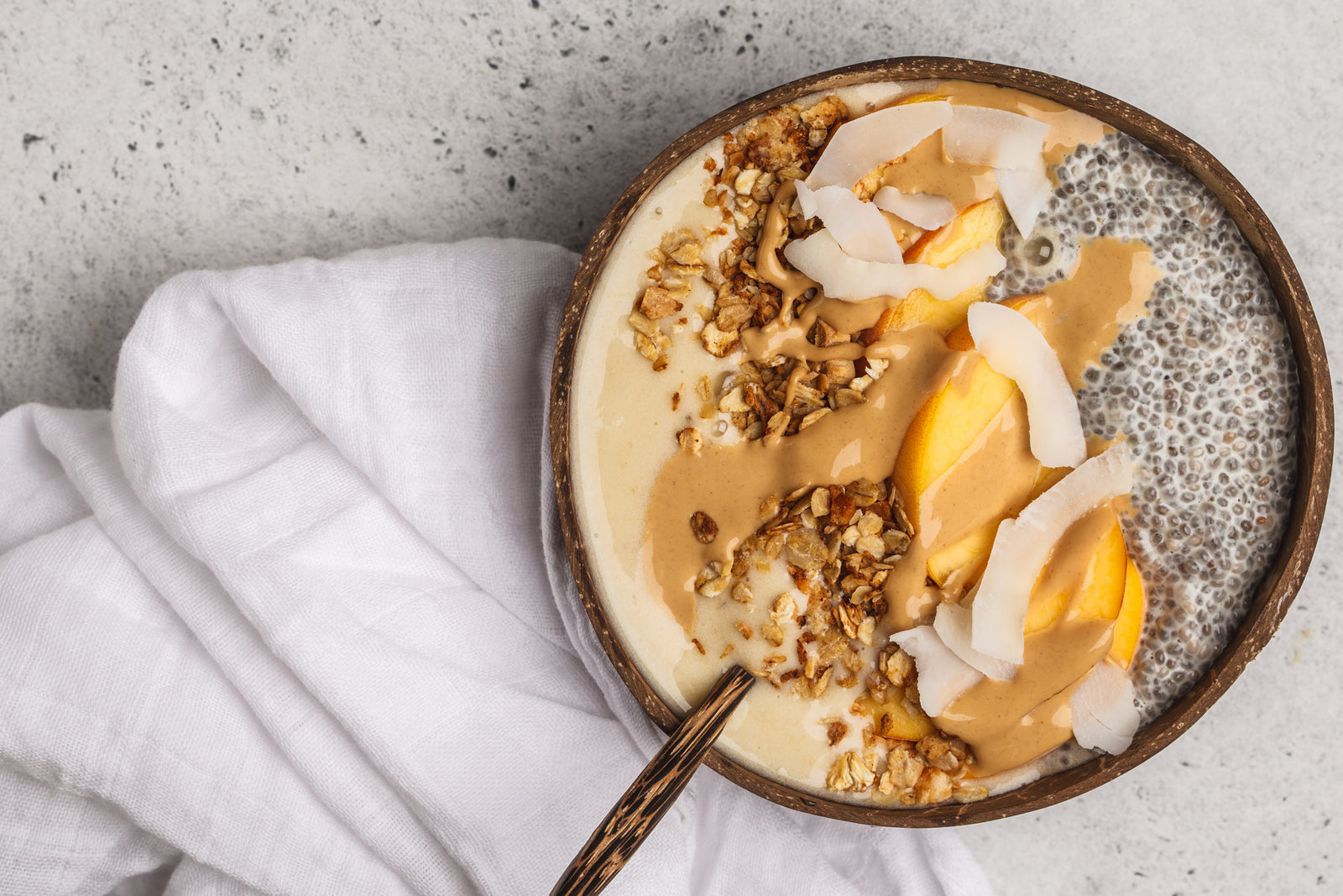 top view of a wooden bowl filled with pineapple chia granola topped with chia seeds, slices of mango, rice flakes, and coconut chips with a wooden spoon  and a white cloth on the side on top of a concrete table 