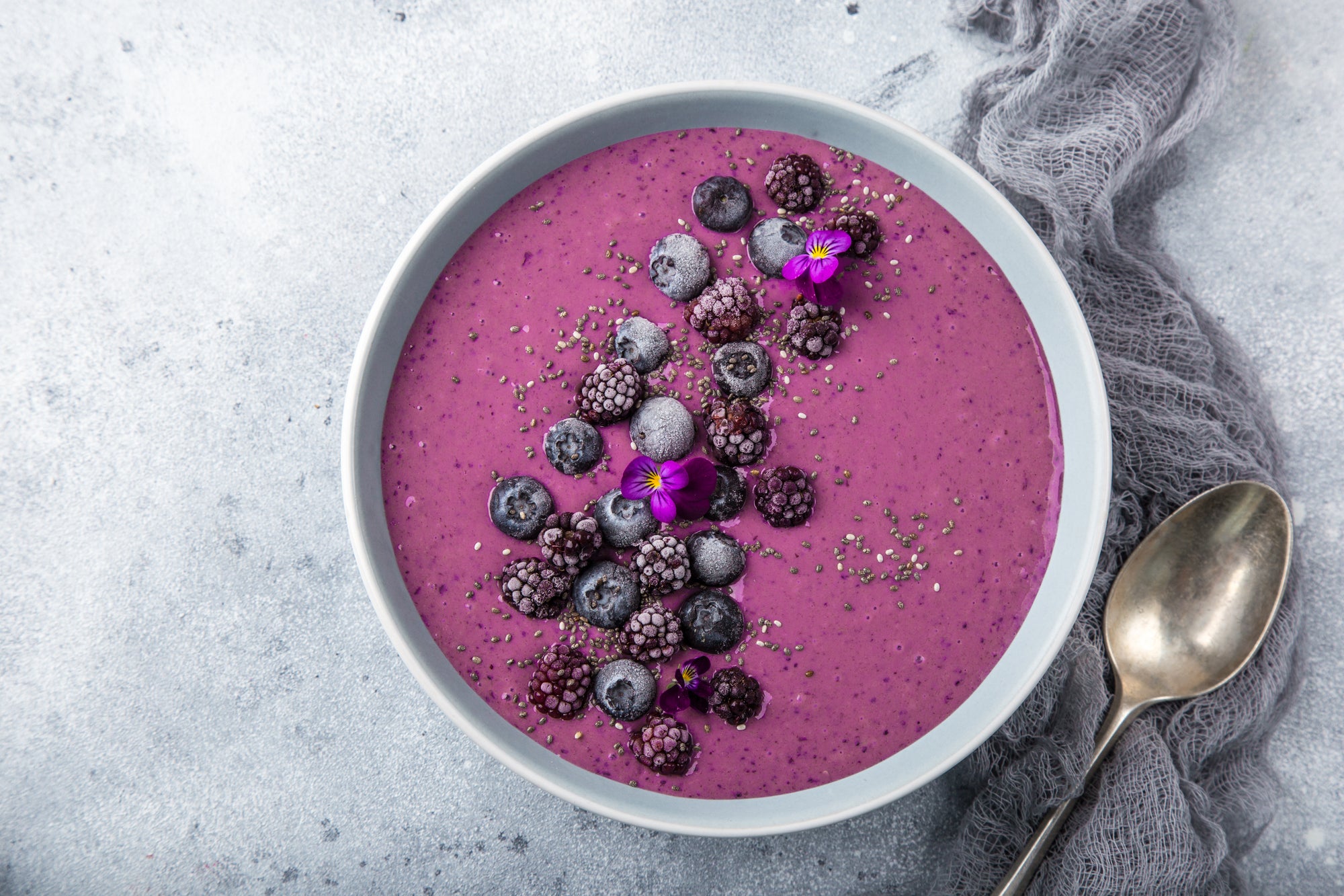 top view of a bowl of powerphenol purple smoothie topped with berries and chia seeds on top of a clothing with a spoon on the side on top of a concrete table