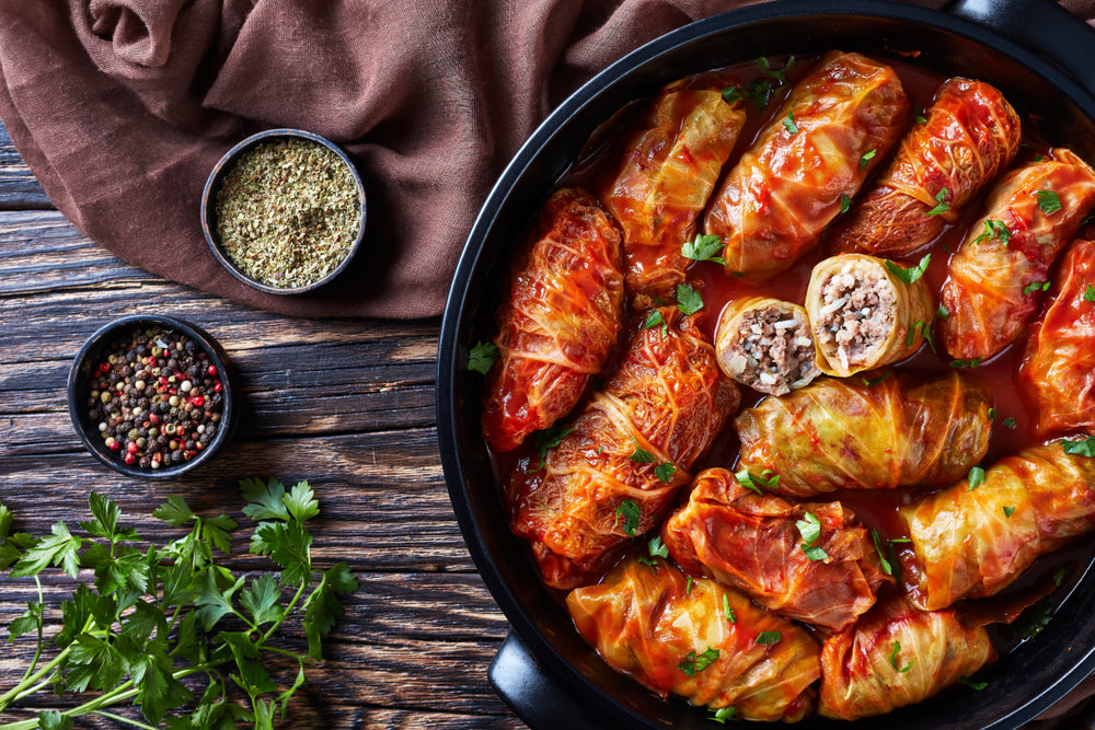 top view of barley stuffed cabbage rolls with brown cloth and small bowls  with peppercorn and dried basil and parsley on the side on top of a wooden table.